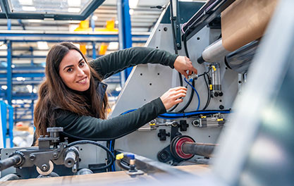 Eine Person arbeitet an einer großen CNC-Industrieanlage und justiert Kabel oder Bauteile. Die Umgebung ist eine helle Produktionshalle mit technischen Anlagen.