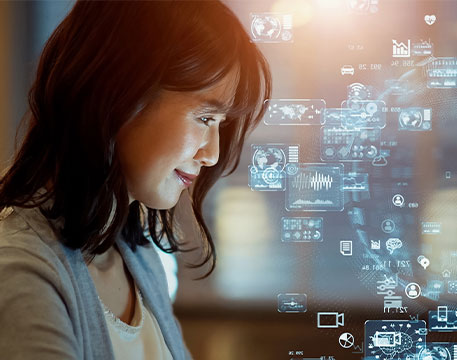 Eine Frau sitzt vor einem Bildschirm und betrachtet eine digitale Benutzeroberfläche, auf der schwebende Symbole wie Diagramme, Datenströme, Kommunikations- und Technologiegrafiken eingeblendet sind.