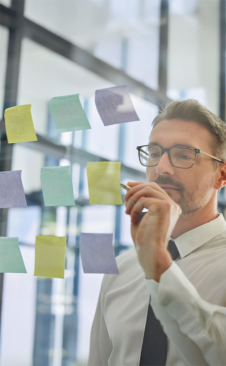 Eine Person steht vor einer Glaswand mit vielen farbigen Haftnotizzetteln. Sie hält einen Stift in der Hand und betrachtet die Notizen konzentriert, als Teil eines Planungs- oder Organisationsprozesses.