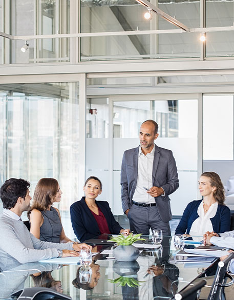 Mehrere Personen sitzen um einen Konferenztisch, während eine stehende Person spricht. Alle sind in ein geschäftliches Gespräch vertieft, umgeben von Glaswänden und hellen Büroflächen.