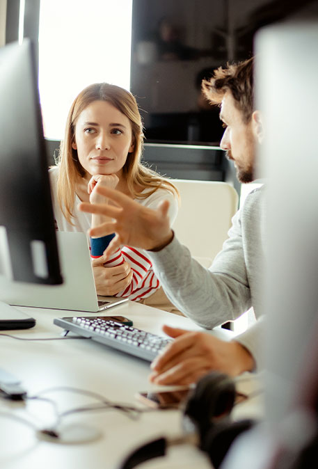 Zwei Personen sitzen an Schreibtischen mit mehreren Monitoren. Eine Person erklärt etwas gestikulierend, während die andere aufmerksam zuhört. Der Arbeitsplatz ist modern und hell gestaltet.